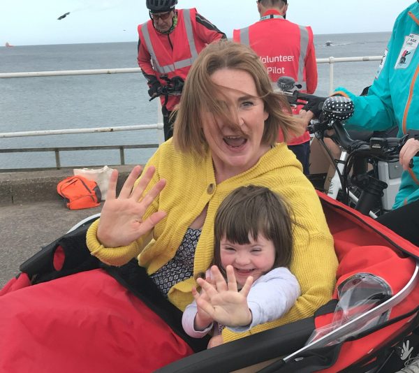 A woman with a yellow jumper is waving and sitting next to a child who is also waving, they are both sitting in a tri-shaw.